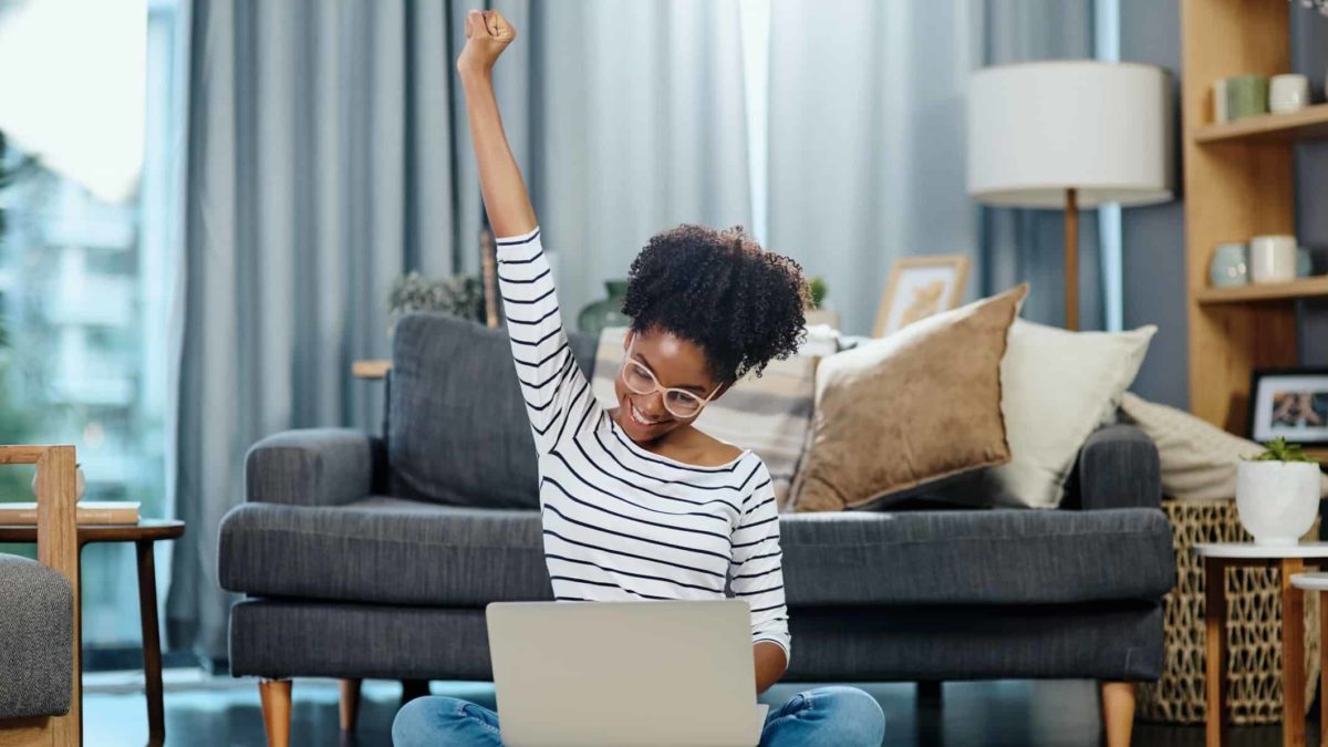A woman sitting in her lounge room punches the air in a gesture of success, having seen the rising IAG share price on her laptop