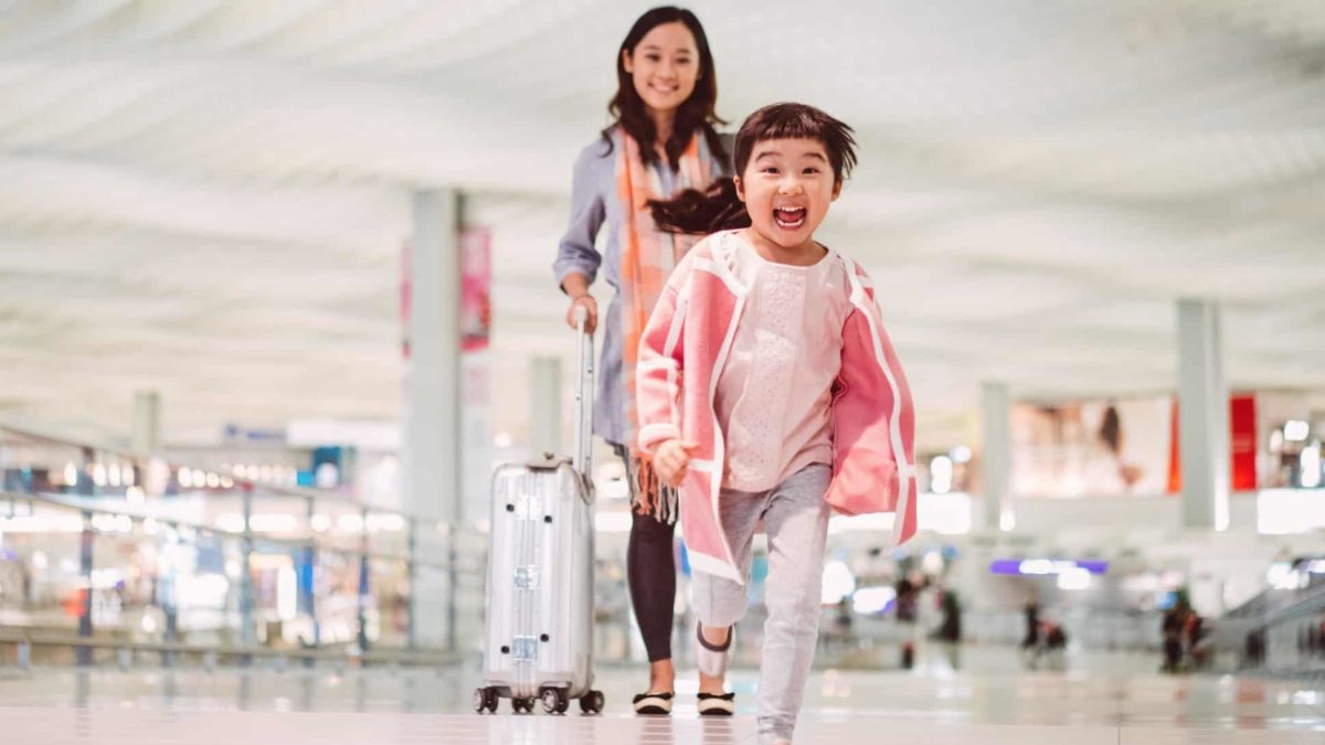 Smiling child runsa towards camera as mum stands beside them with a suitcase at airport.