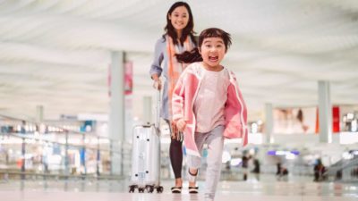 Smiling child runsa towards camera as mum stands beside them with a suitcase at airport.
