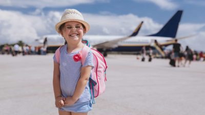 Young smiling girl stands on the tarmac at an airport with a plane in the background.