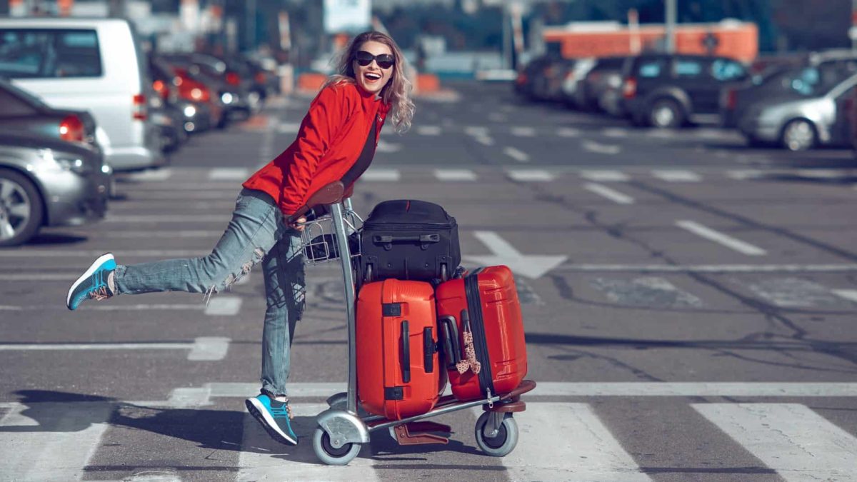 Woman in red smiles as she pushes trolley with suitcases across the road at an airport.