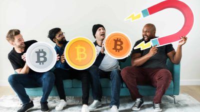 Three men sit in a row holding giant bitcoins while the fourth wields a huge magnet