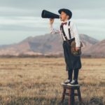 An old-fashioned news boy stands on a stool and yells through a microphone in an open field.