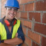 A young male builder with his arms crossed leans against a brick wall and smiles at the camera as the Brickworks share price climbs today