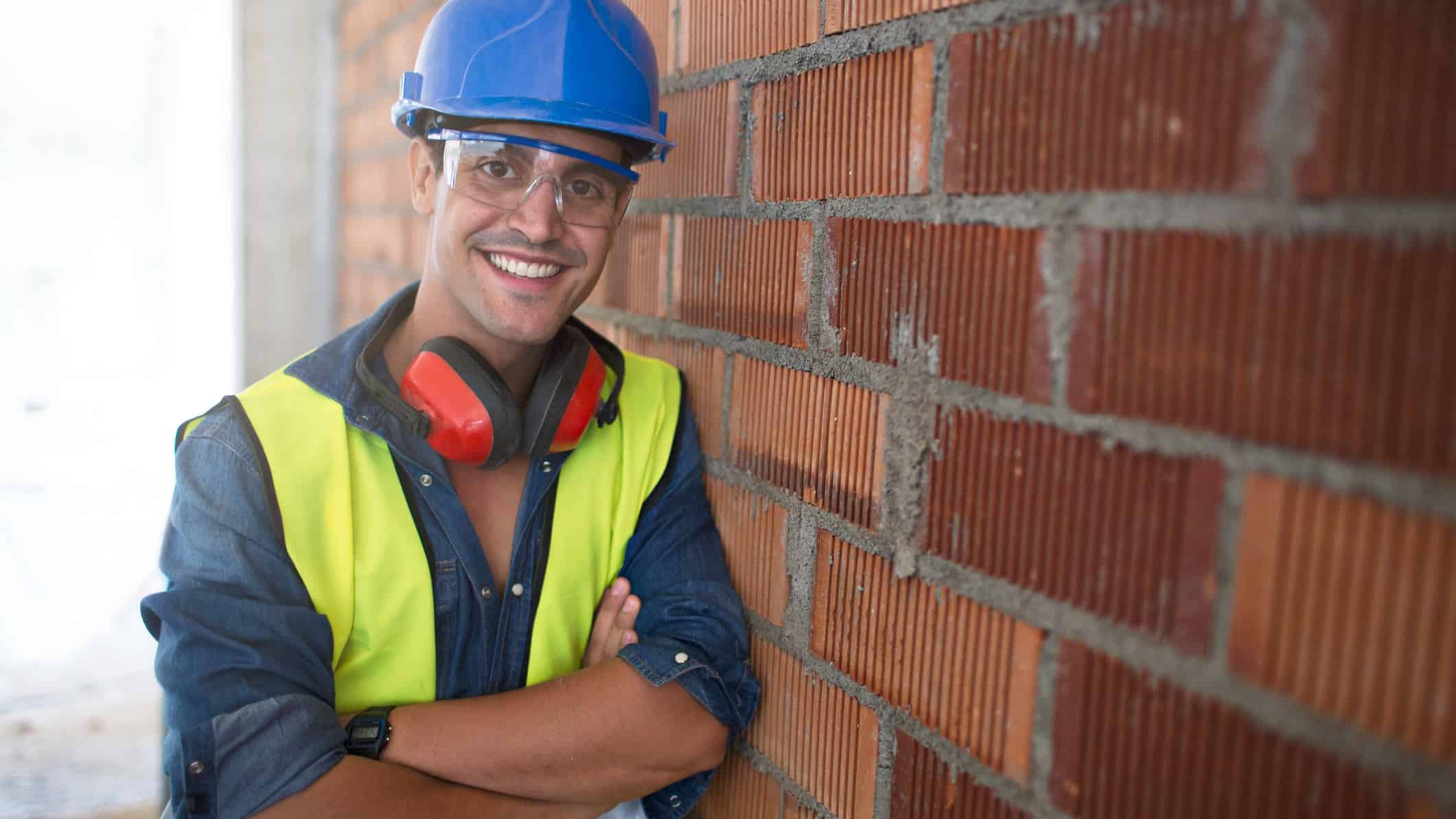 A young male builder with his arms crossed leans against a brick wall and smiles at the camera as the Adbri share price climbs today