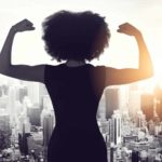 A business woman flexes her muscles overlooking a city scape below.