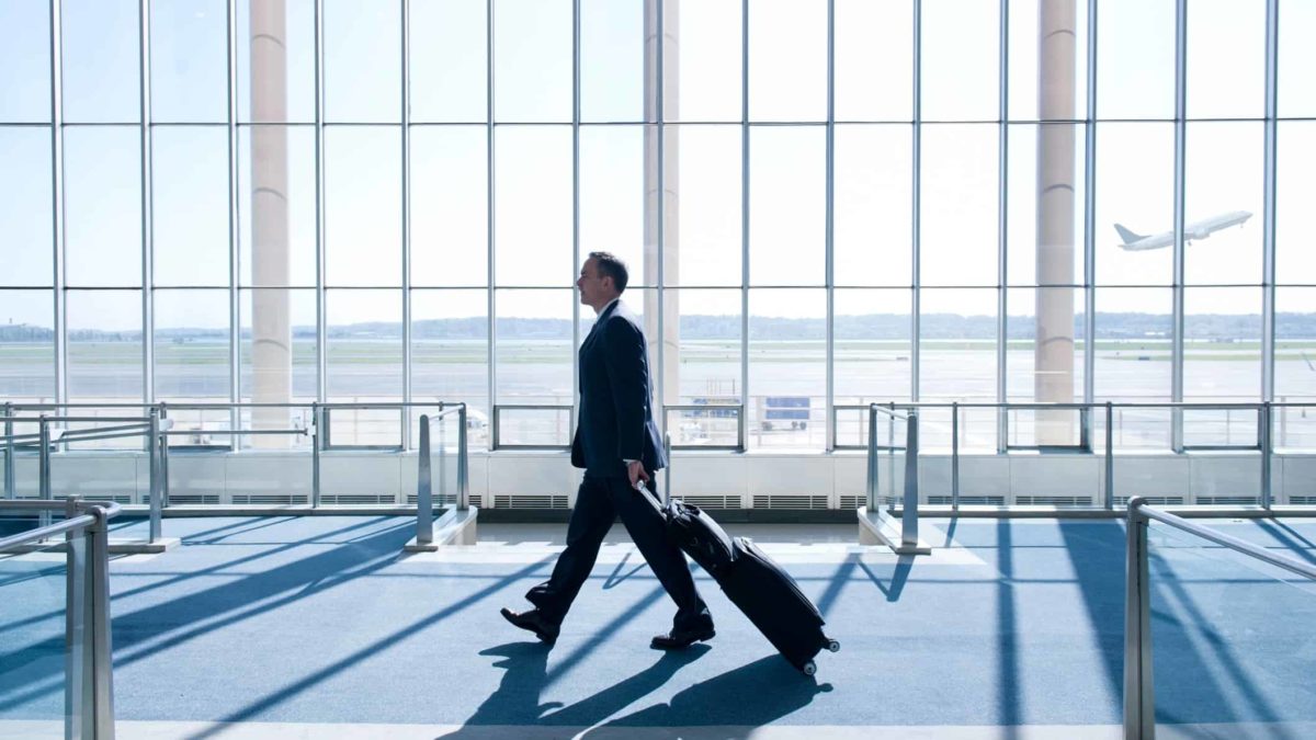 A man in a dark blue suit walks through an airport past floor-to-ceiling windows with a Qantas plane flying in the distance