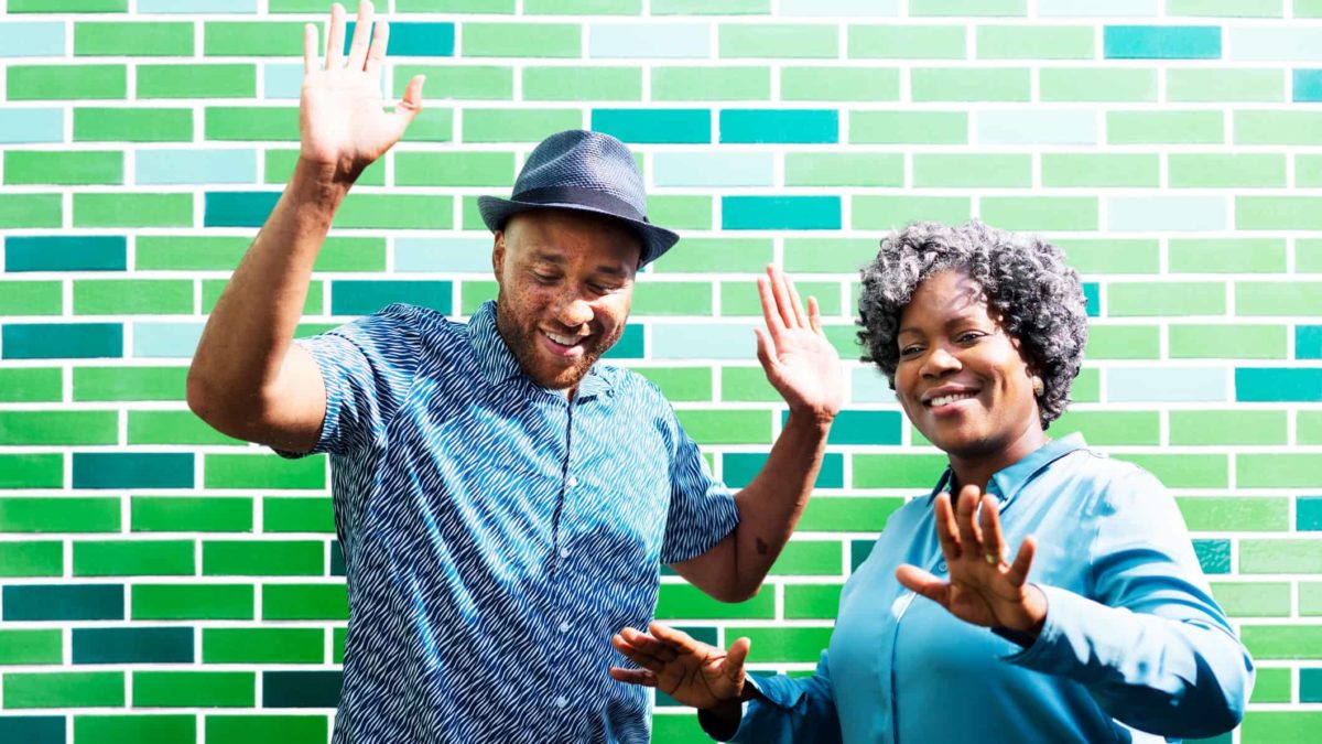 A man and woman put hands in the air as they dance in front of a green brick wall.