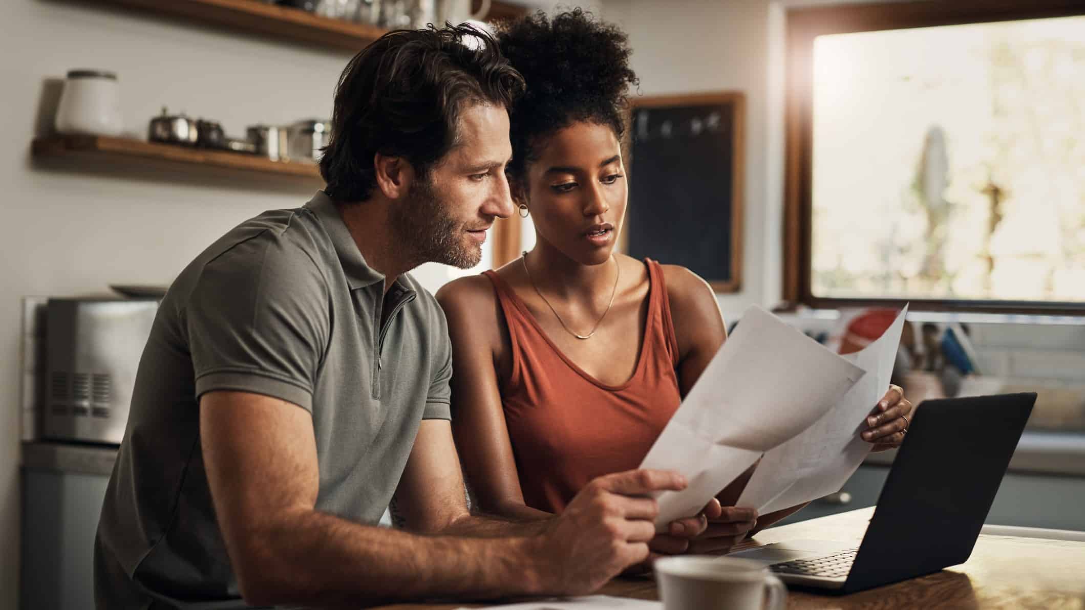 A young couple sits at their kitchen table looking at documents with a laptop open in front of them