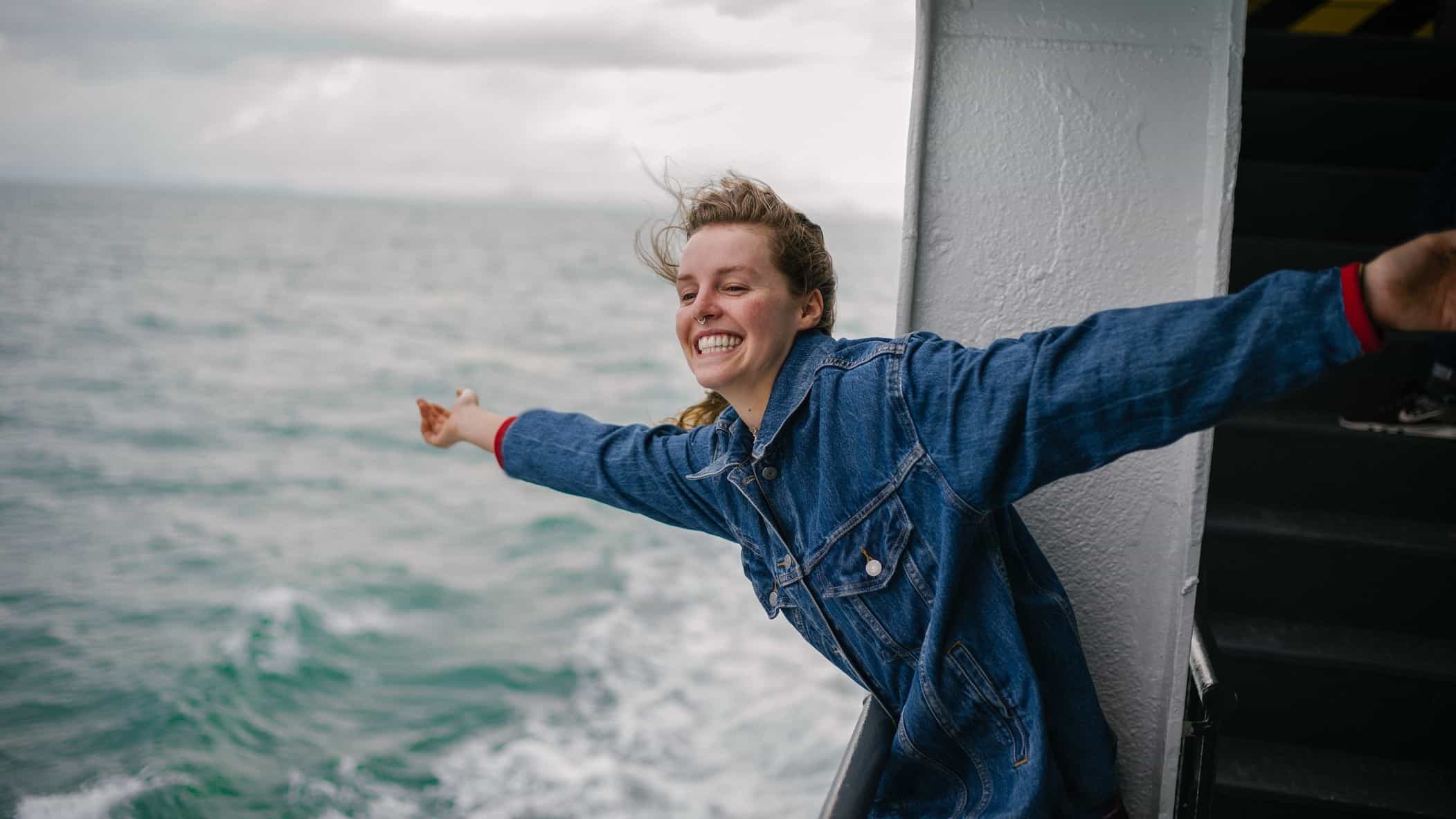 A young female traveller leans over the balcony of her cruise ship room and holds her arms out enjoying the sea air