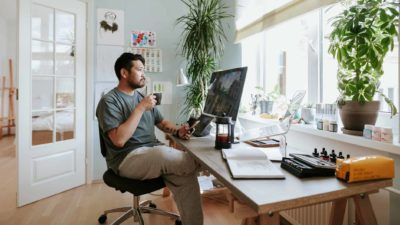 A young man working from home sits at his home office desk holding a cup of tea and looking out the window