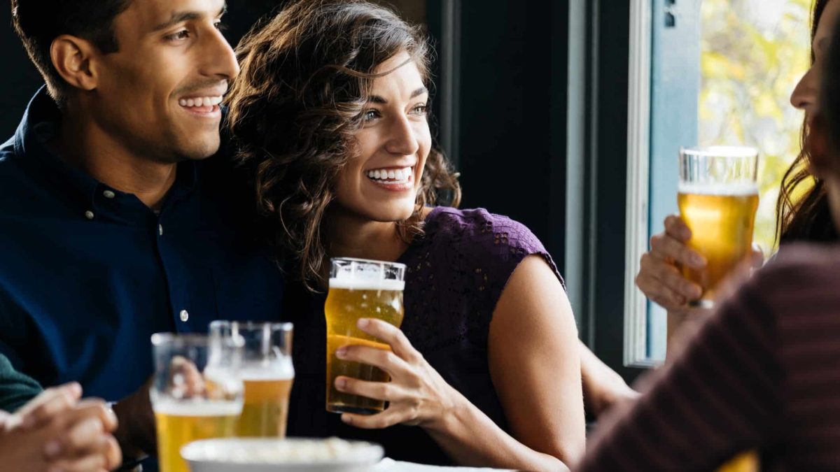 A young couple snuggles together in a pub sitting at a table with friends and laughing