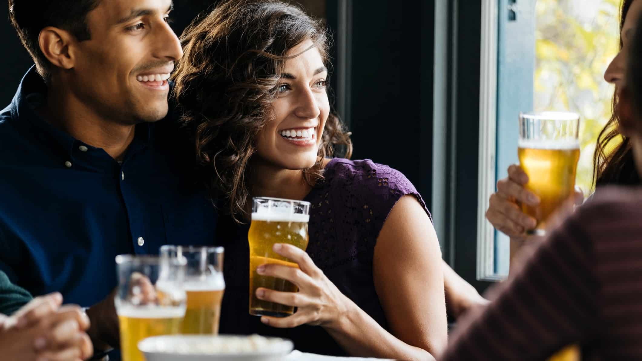 A young couple snuggles together in a pub sitting at a table with friends and laughing