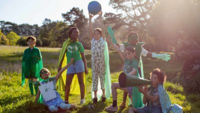 A group of eco warrior children together in nature wear green and capes and hold up a globe of the world..