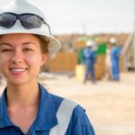 A female worker in a hard hat smiles in an oil field.