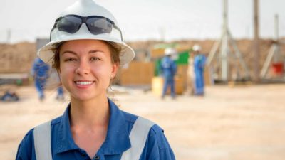 A female worker in a hard hat smiles in an oil field.