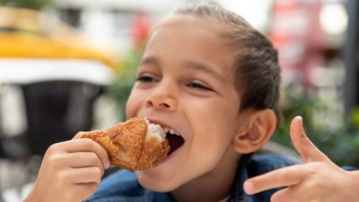 A young boy points and smiles as he eats fried chicken.