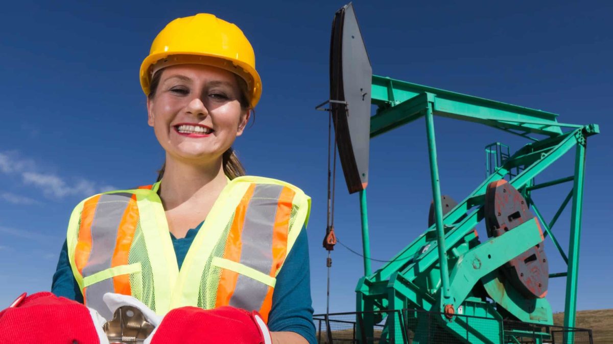 Female oil rig worker wearing high vis vest, red gloves and hardhat smiles at camera with a green painted oil rig in the background