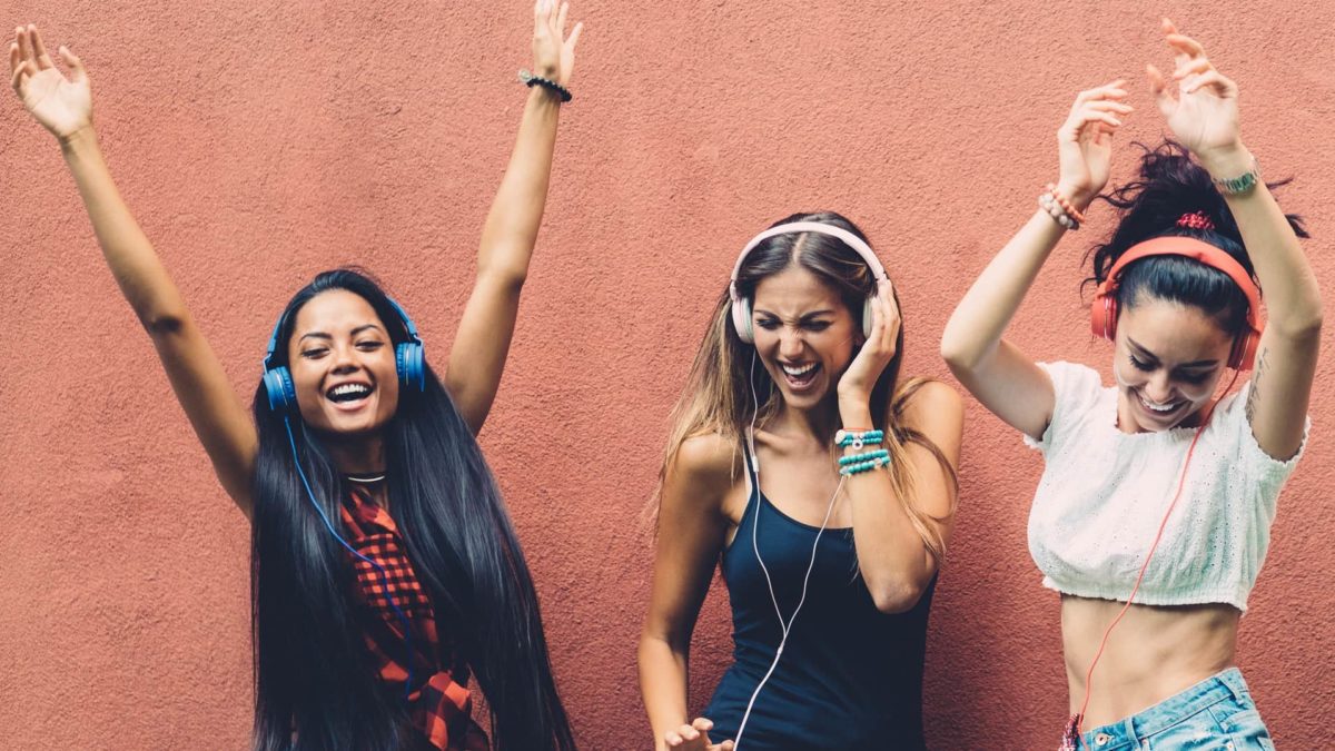 Three happy young women wearing headphones dance to music.