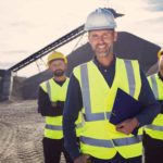 Three mining workers stand proudly in front of a mine smiling because the BHP share price is rising