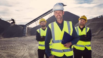 Three mining workers stand proudly in front of a mine smiling because the BHP share price is rising