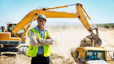 A miner in a hard hat and high visibility vest stands with his arms crossed with a large digging machine in the background.