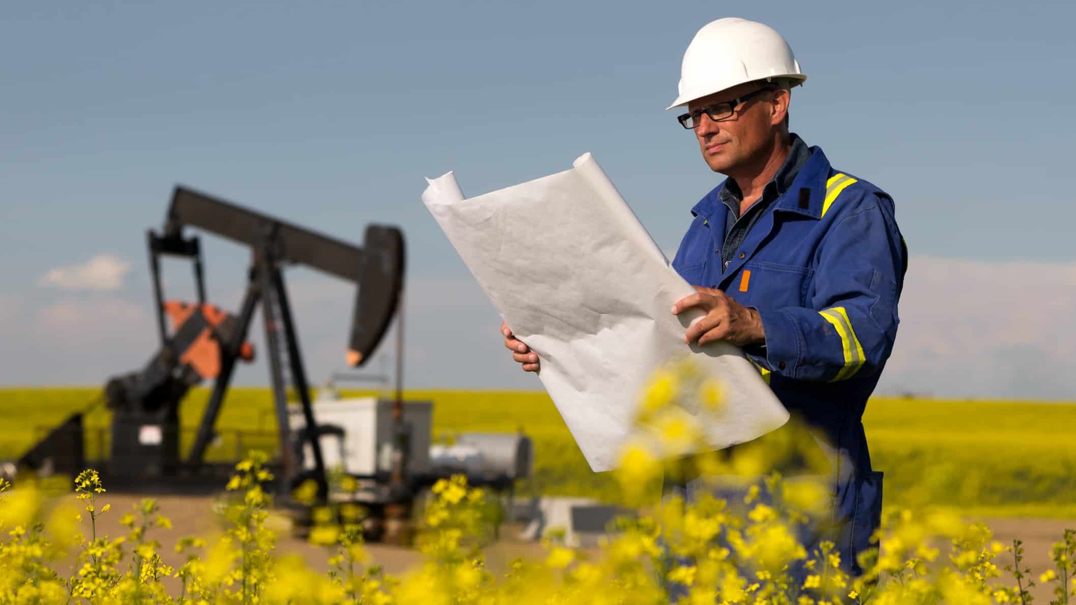 An oil mining worker wearing a hard hat stands in a yellow field looking at blueprints with an oil rig and blue sky in the background