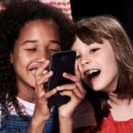 Two girls smile and laugh as they use a mobile phone.
