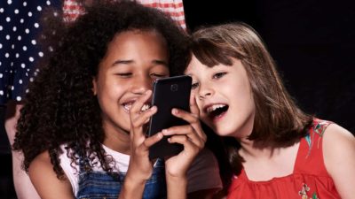 Two girls smile and laugh as they use a mobile phone.