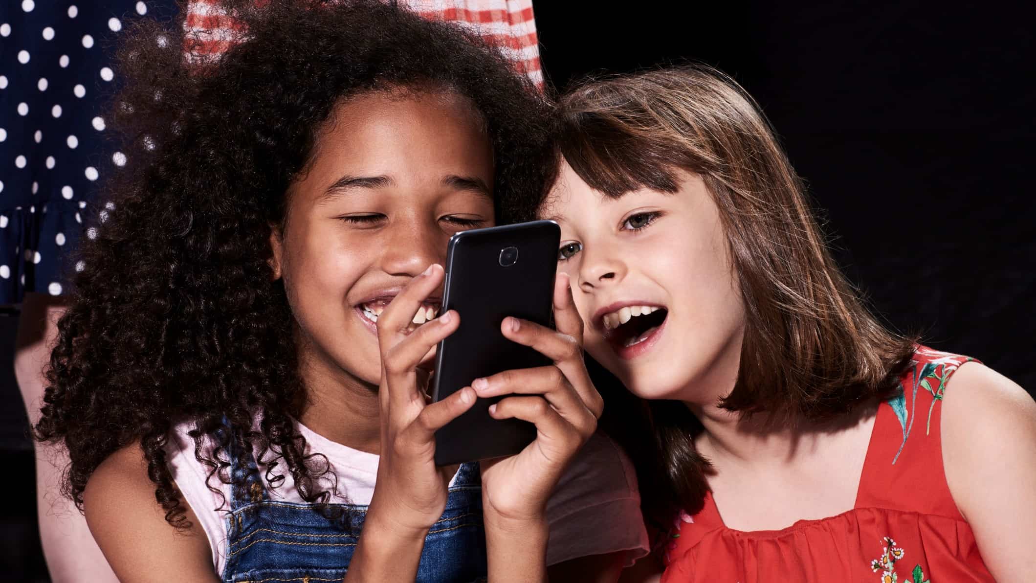 Two girls smile and laugh as they use a mobile phone.