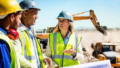 Mining workers in high vis vests and hard hats discuss plans for the mining site they are at as heavy equipment moves earth behind them, representing opportunities among ASX 200 shares as nominated by top broker Macquarie