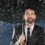 Man standing with an umbrella over his head with a sad face whilst it rains.