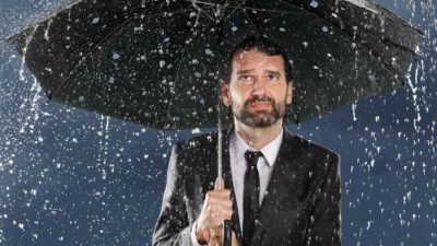 Man standing with an umbrella over his head with a sad face whilst it rains.