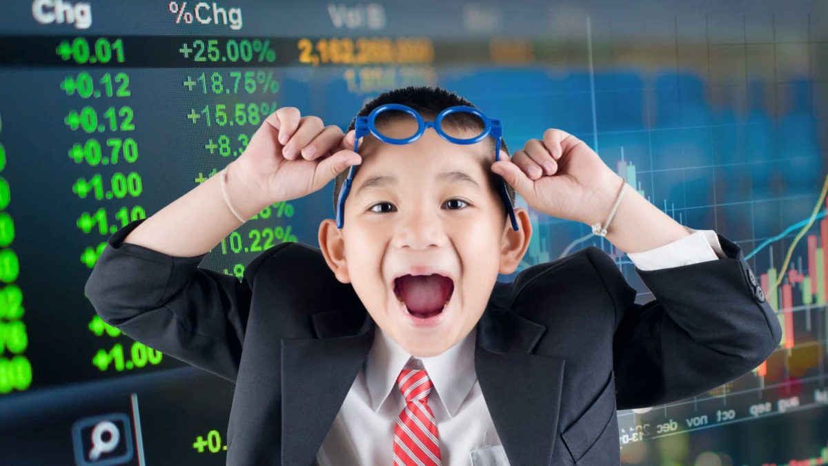 A young boy in a business suit lifts his glasses above his eyes and gives a big wide mouthed smile to the camera with a stock market board in the background.