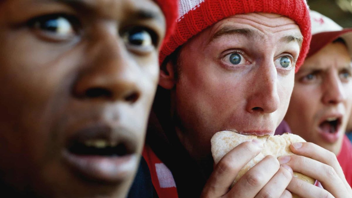 Football supporters at match, one holding hambuger, in a close-up shot.