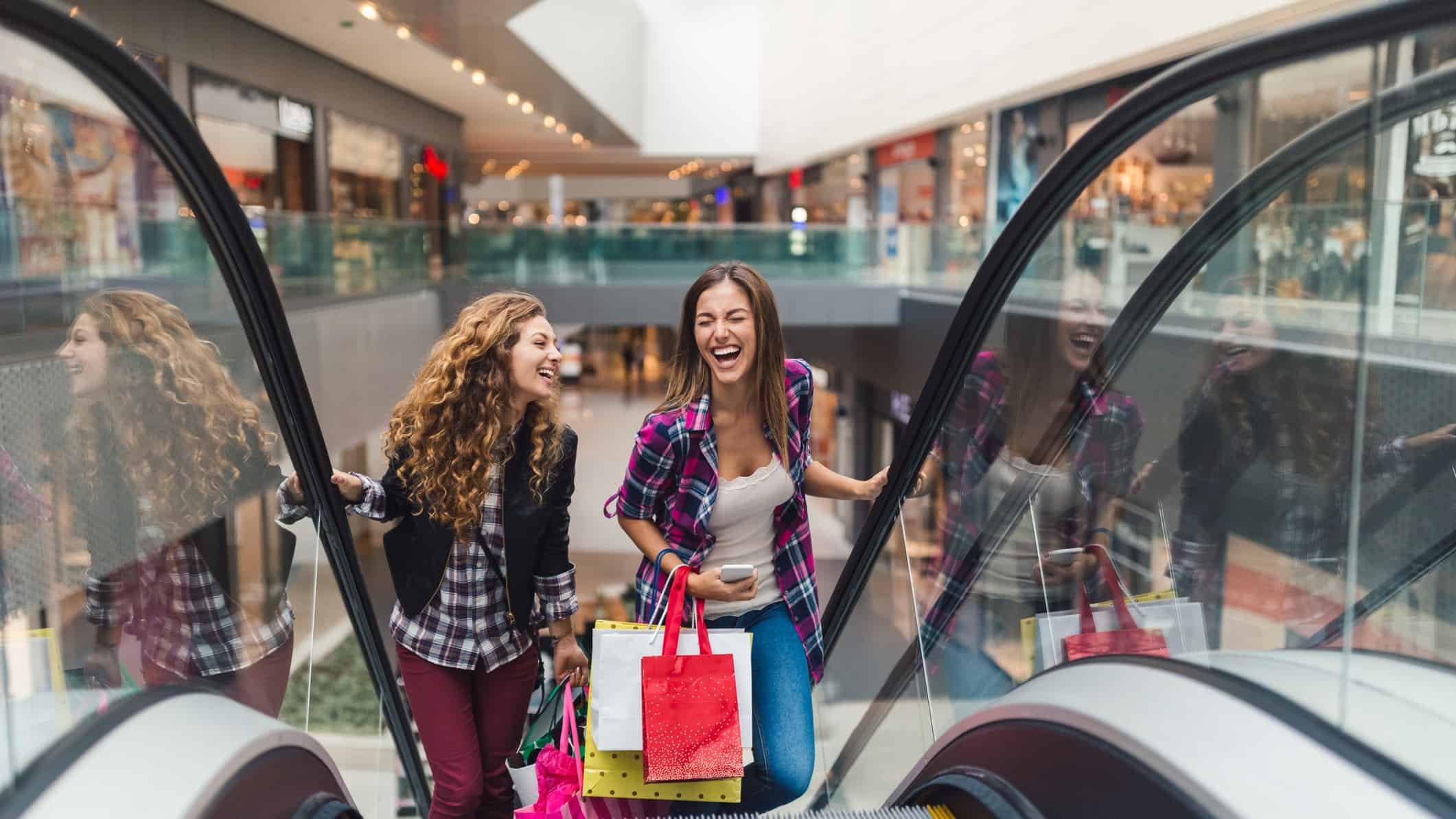 Two laughing young women holding shopping bags ride an escalator up to another level in a Scentre Group shopping centre