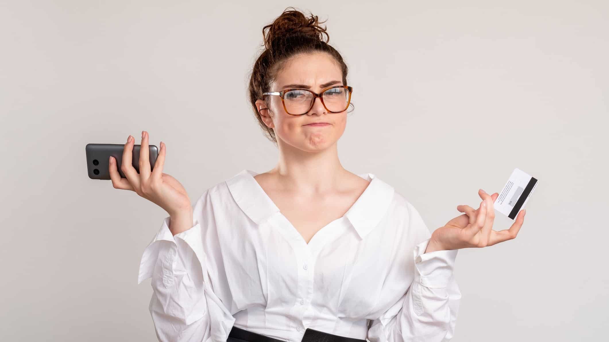 Puzzled female client shrugging with credit card phone isolated on light background.