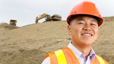 A man in a hard hat stands in the foreground of a large mound of earth with heavy moving equipment on top.