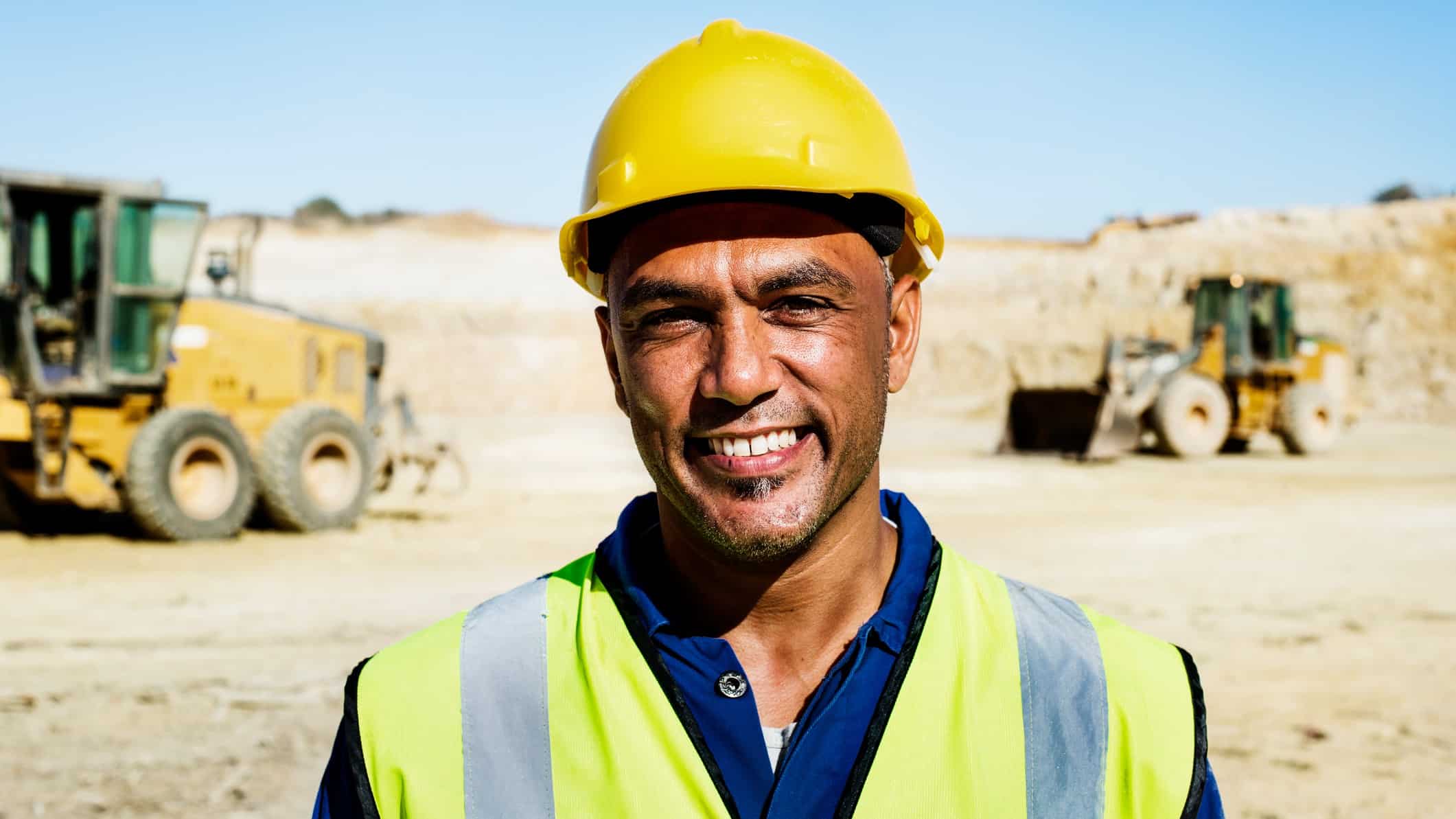 a miner wearing a hard hat smiles as he stands in front of heavy earth moving equipment on a barren mine site.