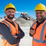 Two smiling men in high visibility vests and yellow hardhats stand side by side with a large mound of earth and mining equipment behind them smiling as the Carnaby Resources share price rises today