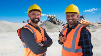 Two smiling men in high visibility vests and yellow hardhats stand side by side with a large mound of earth and mining equipment behind them smiling as the Carnaby Resources share price rises today