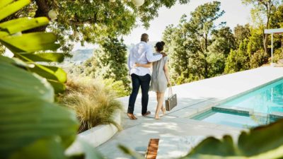 A relaxed couple arm in arm stroll alongside a hotel pool with beautiful greenery around them feeling excited about their holiday travel