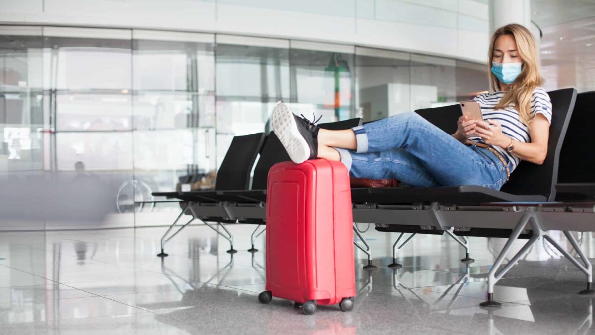 A young woman with a mask sits in an airport with her feet resting on top of her red suitcase while she looks at her phone