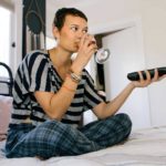 Woman sits cross legged on bed drinking a glassing of wine and holdaing TV remote control.