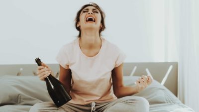 CA woman sits on her bed wailing and crying with a wine bottle in one hand and a glass in the other.