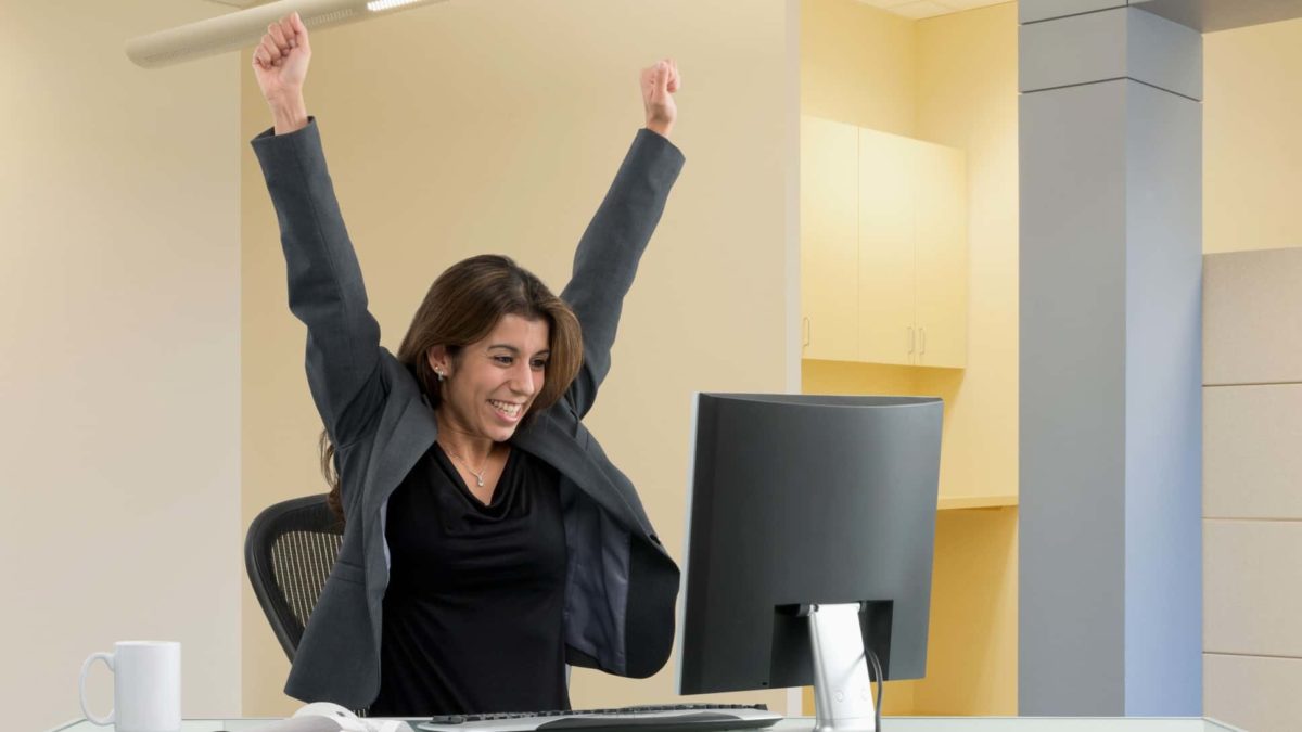 A happy woman in an office puts her hands in the air as if to celebrate while looking at computer.