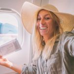 A smiling woman in a hat holding a ticket takes selfie inside a Qantas plane next to the window.