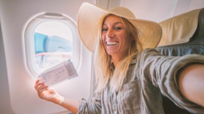 A smiling woman in a hat holding a ticket takes selfie inside a Qantas plane next to the window.