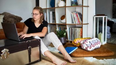 A young woman wearing glasses and casual clothes working from home sits on the floor and types on her laptop that is on a coffee table next to her
