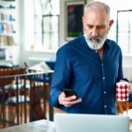 A middle-aged man working from home looks at his mobile phone with a laptop open on the table in front of him.